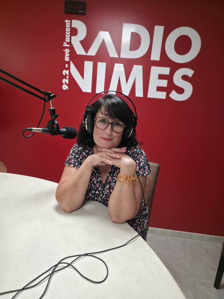 Femme souriante portant des écouteurs, assise devant un microphone dans un studio de radio, avec un mur rouge sur lequel est écrit 'RADIO NIMES'.